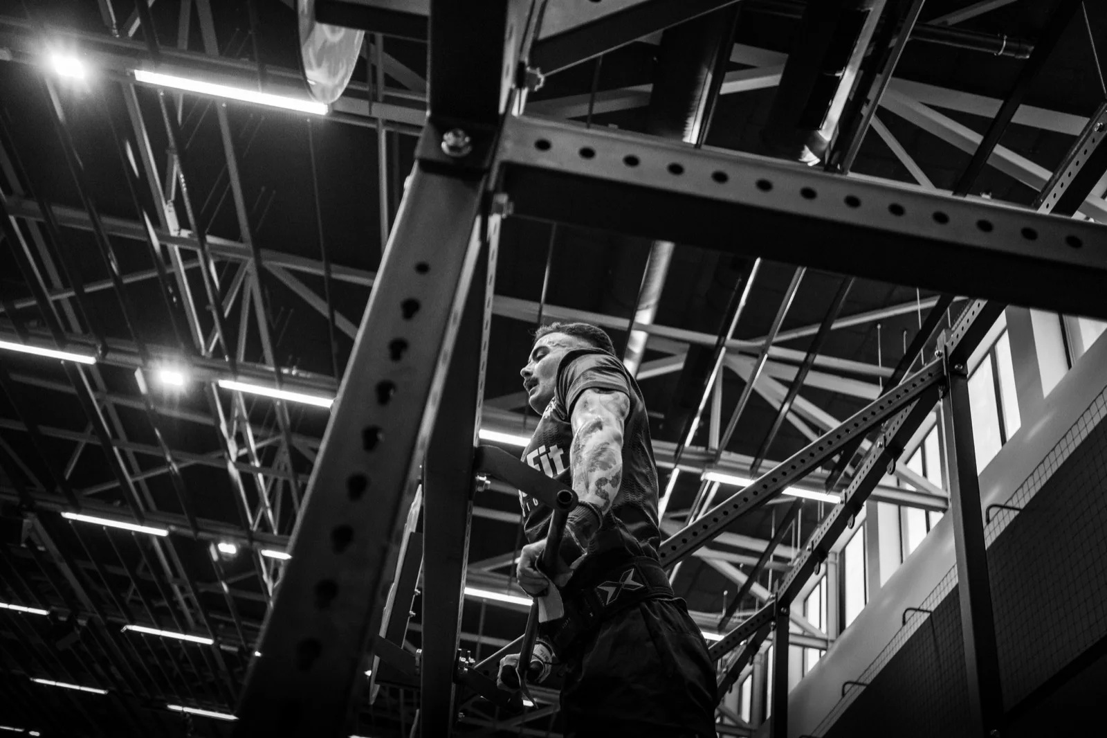 Low-angle black-and-white photograph of an EMT competitor climbing a competition rig, with the hall ceiling structure in the background